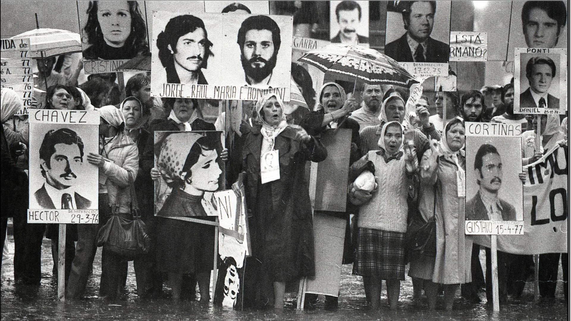Madres de plaza de mayo sosteniendo pancartas con fotografías de sus hijos desaparecidos bajo la lluvia.